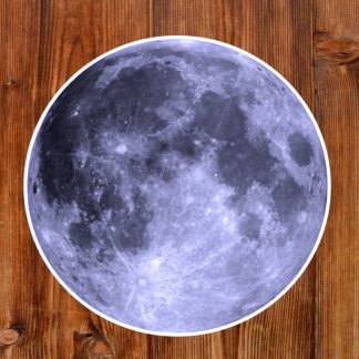 Gothic sticker of a realistic full moon image with detailed craters sits on a textured wooden surface. The contrast highlights the moon's rugged beauty against the warm wood.