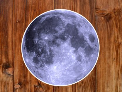 Gothic sticker of a realistic full moon image with detailed craters sits on a textured wooden surface. The contrast highlights the moon's rugged beauty against the warm wood.
