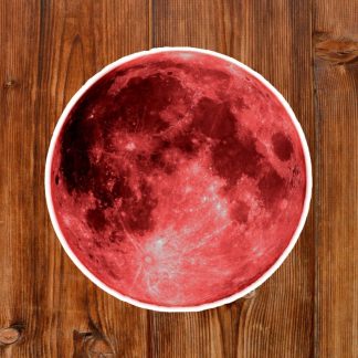 Gothic round sticker of a vibrant blood red moon with a detailed lunar pattern on a rustic wooden surface. The visual contrast highlights the moon's craters and textures.