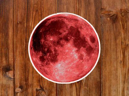 Gothic round sticker of a vibrant blood red moon with a detailed lunar pattern on a rustic wooden surface. The visual contrast highlights the moon's craters and textures.