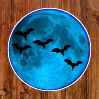 Gothic sticker features bright blue moon with five silhouetted bats flying across it, set against a warm wooden background, creating a spooky and mysterious atmosphere.