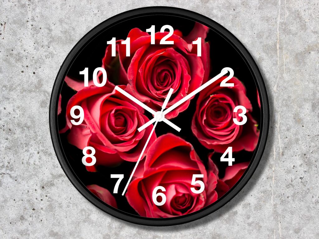 A round wall clock featuring vibrant red roses on a black background. White hour numbers and hands contrast against the vivid floral design, creating an elegant, romantic tone.