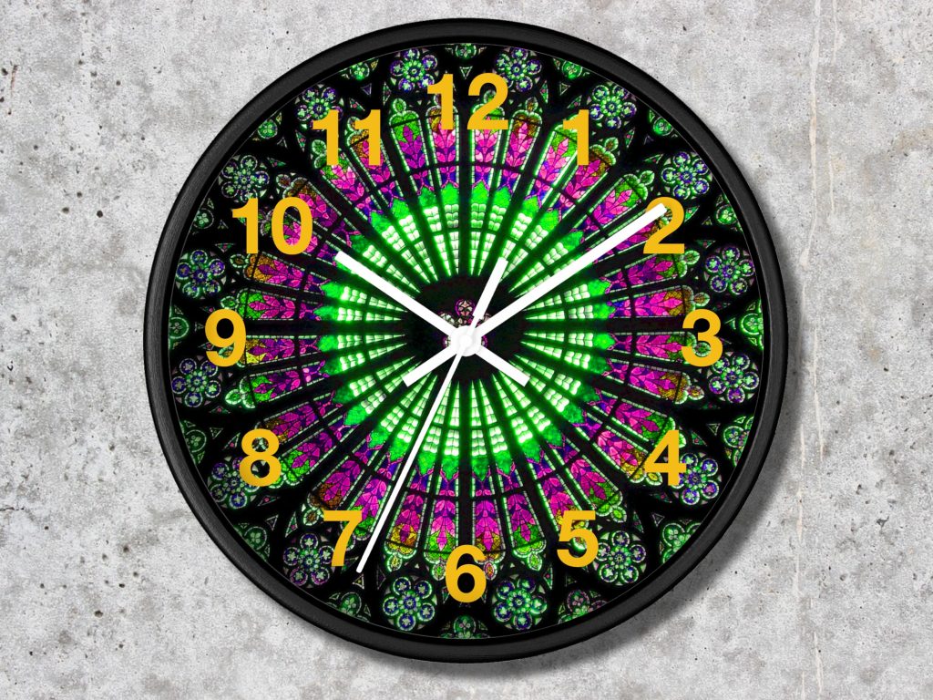 A colorful clock in a gothic style features a vibrant stained glass pattern with green and pink hues. Yellow numbers contrast against the dark frame, evoking a dynamic and artistic feel.
