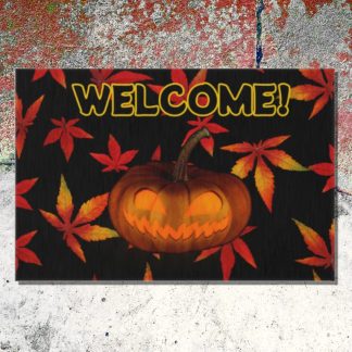 A Halloween-themed welcome floor mat features a carved pumpkin with a mischievous grin, surrounded by red and orange autumn leaves, set against a textured, weathered cement surface.