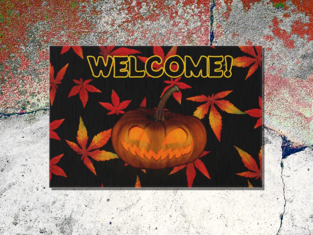 A Halloween-themed welcome floor mat features a carved pumpkin with a mischievous grin, surrounded by red and orange autumn leaves, set against a textured, weathered cement surface.