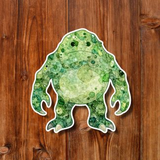 Green watercolor frog-shaped creature monster sticker with white outline rests on rustic wooden planks, its mottled texture creating a playful, whimsical mood.