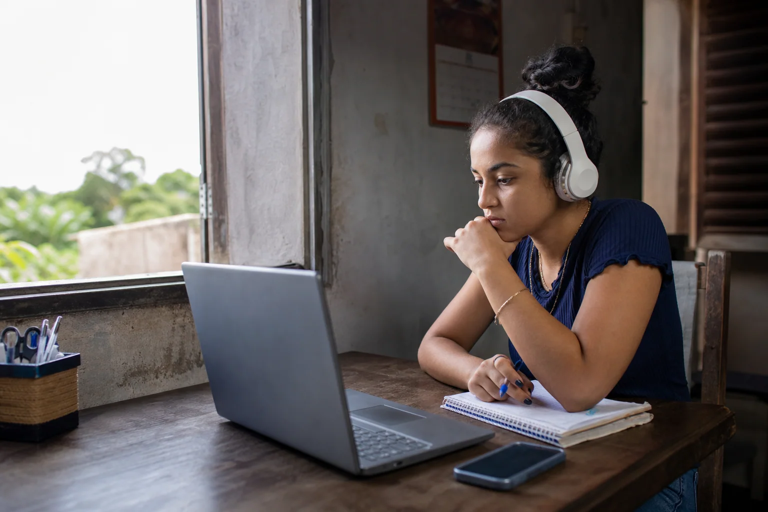 Jovem em casa analisa no notebook uma vaga de curso gratuito de inglês, com caderno e celular sobre a mesa.