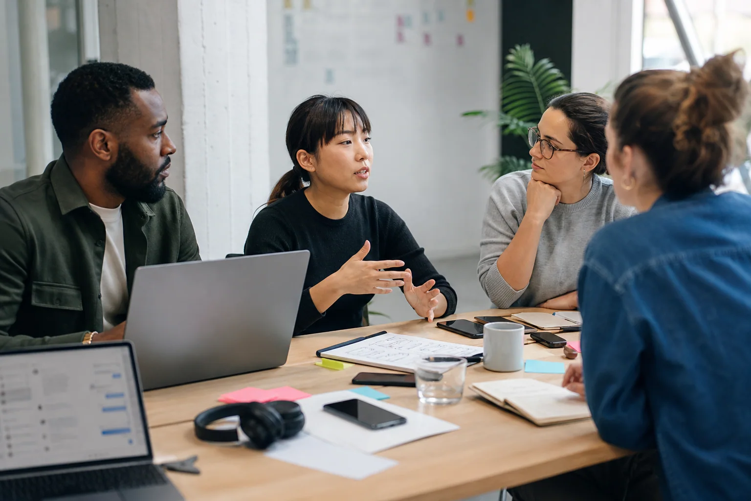 Profissionais em ambiente de trabalho colaborativo conversam de forma natural em uma mesa de escritório, com notebooks e anotações discretas, em clima de comunicação clara e alinhamento sem exagero.