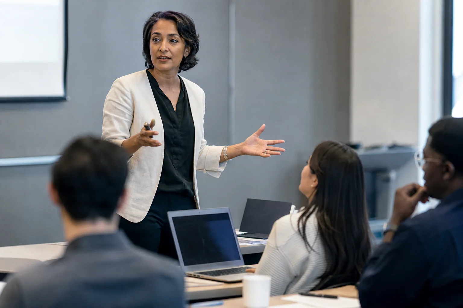 Professor ou pesquisadora conduzindo uma aula ou apresentação em inglês para um pequeno grupo em ambiente acadêmico, com gesto explicativo e público atento.