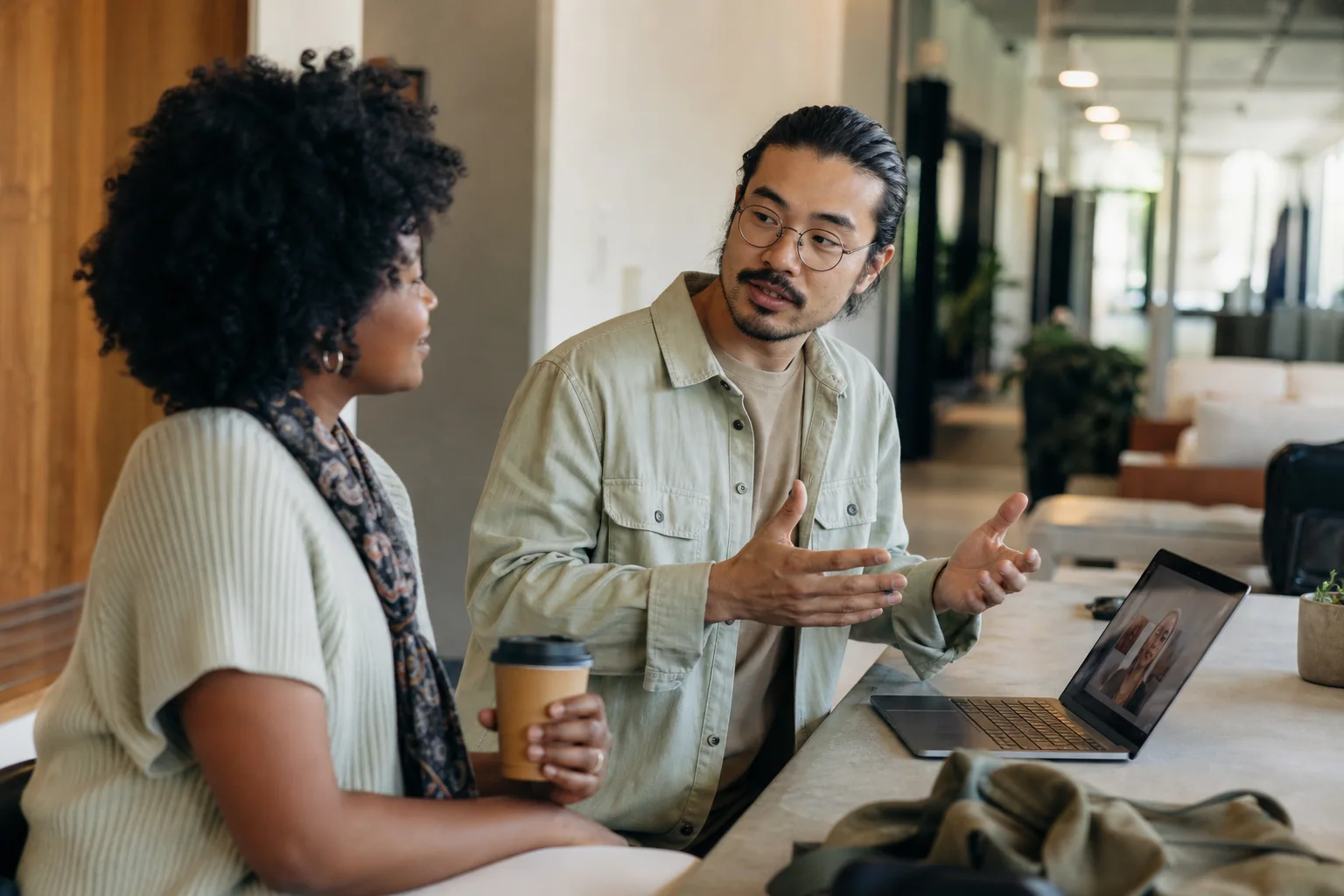 Duas pessoas conversam de forma natural em um espaço híbrido de trabalho e café, com um laptop aberto ao fundo, sugerindo small talk em inglês em situações do dia a dia.