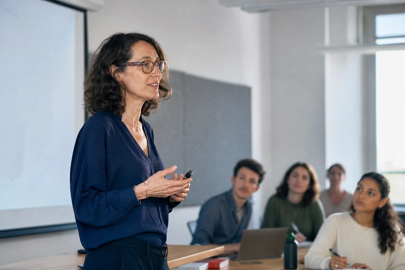 Professora conduzindo uma explicação em inglês para uma turma universitária, em ambiente acadêmico claro e contemporâneo.