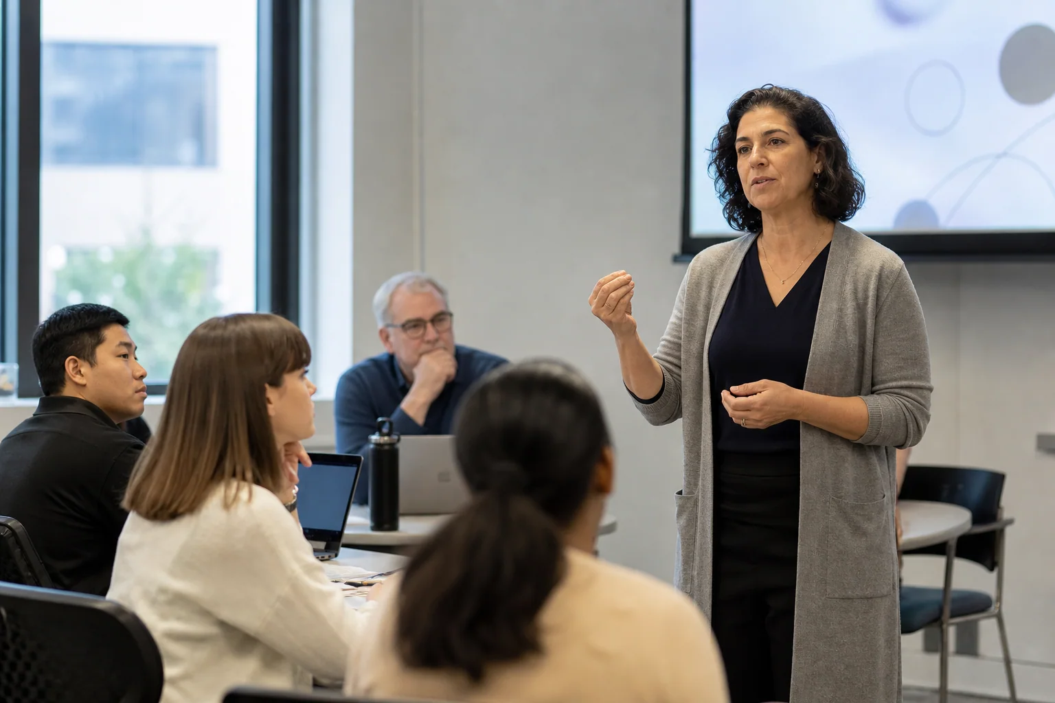 Professora conduz uma aula ou apresentação acadêmica em inglês para um pequeno grupo em ambiente universitário contemporâneo.