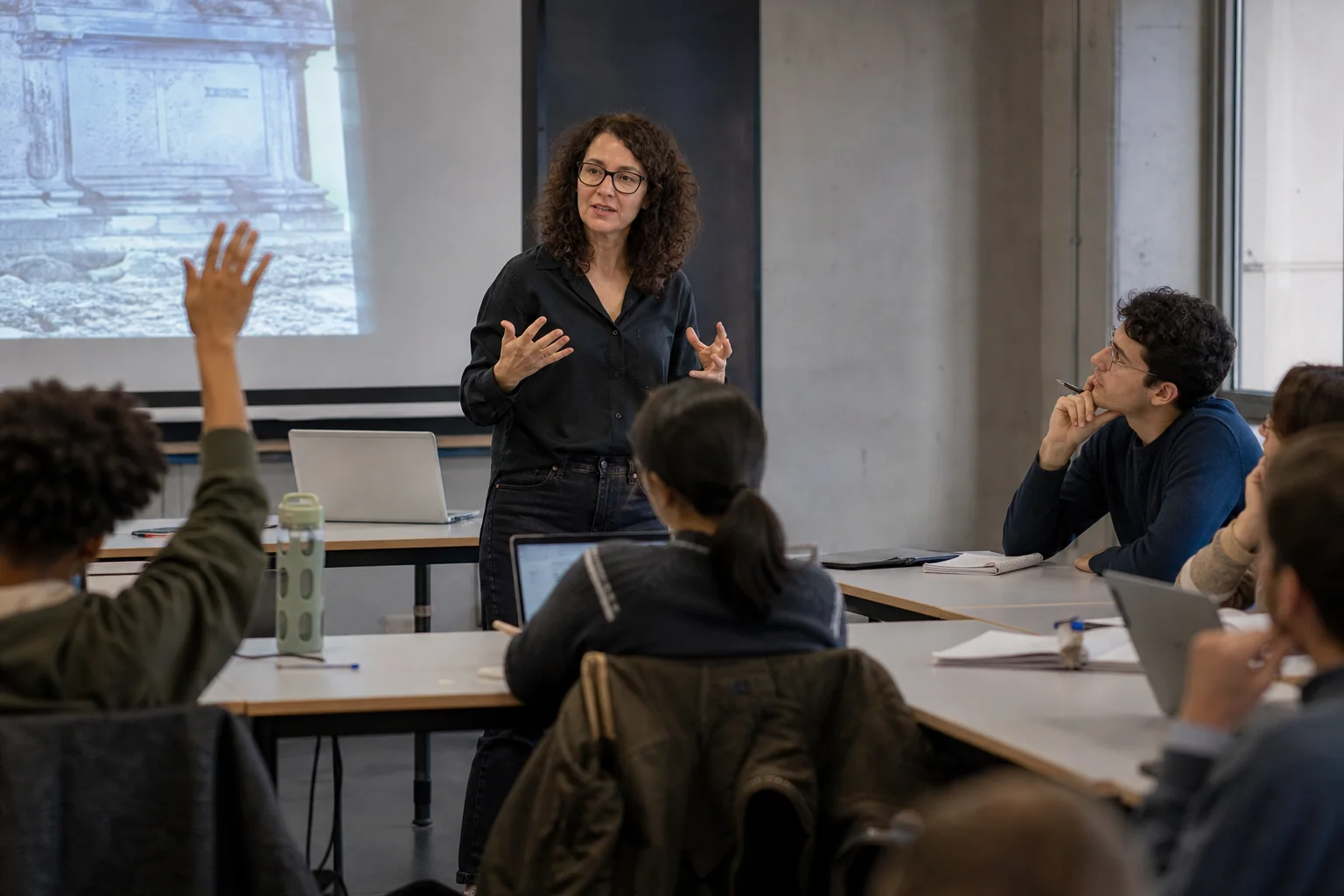 Professora ou pesquisadora explica um tema e responde a uma pergunta em uma sala universitária, com alunos atentos em um ambiente acadêmico contemporâneo.