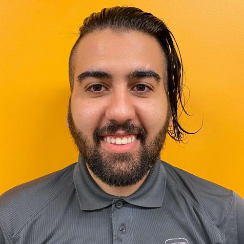 Corey standing in front of a yellow wall smiling wearing a gray TWO MEN AND A TRUCK polo