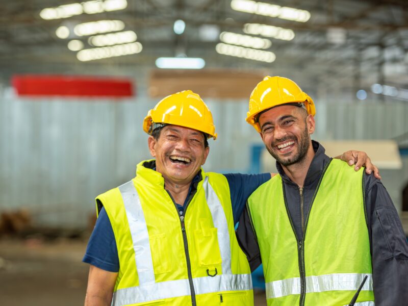 Two happy factory workers wearing hard hats and safety vests standing with arms crossed.