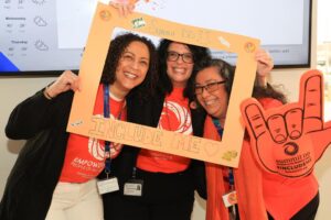 3 women wearing orange, smiling while holding a hand-made Inclusion Day photo frame for Summit DD's Inclusion Day