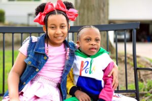 Young girl with bow sitting with her arm around her younger brother on a park bench