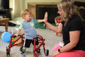 Little boy in a stander giving high five to developmental specialist sitting next to him