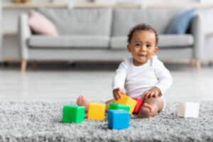 Infant Child Playing With Stacking Building Blocks At Home, Adorable Little Baby Sitting On Carpet In Living Room And Looking At Camera,