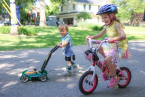 Toddler walking with play lawn mower next to little girl riding a small 2-wheel bicycle