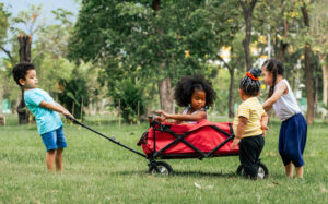 Four cute little children pull and drag a red cart while playing in outdoor green park.
