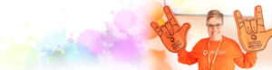 Image of smiling woman wearing orange for inclusion and holding up 2 foam hands in the ASL sign of Love