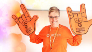 Image of smiling woman wearing orange for inclusion and holding up 2 foam hands in the ASL sign of Love