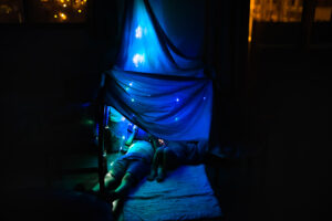 Two young kids lying on the floor of their house under bed sheets lit with light bulbs.