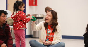 Smiling developmental specialist sits on floor helping small child with green toy