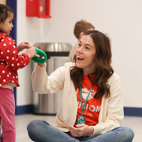 Smiling developmental specialist sits on floor helping small child with green toy