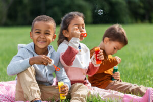 group of children, sitting on a blanket, blowing soap bubbles at park