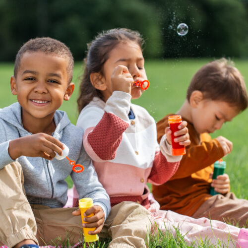 group of children, sitting on a blanket, blowing soap bubbles at park