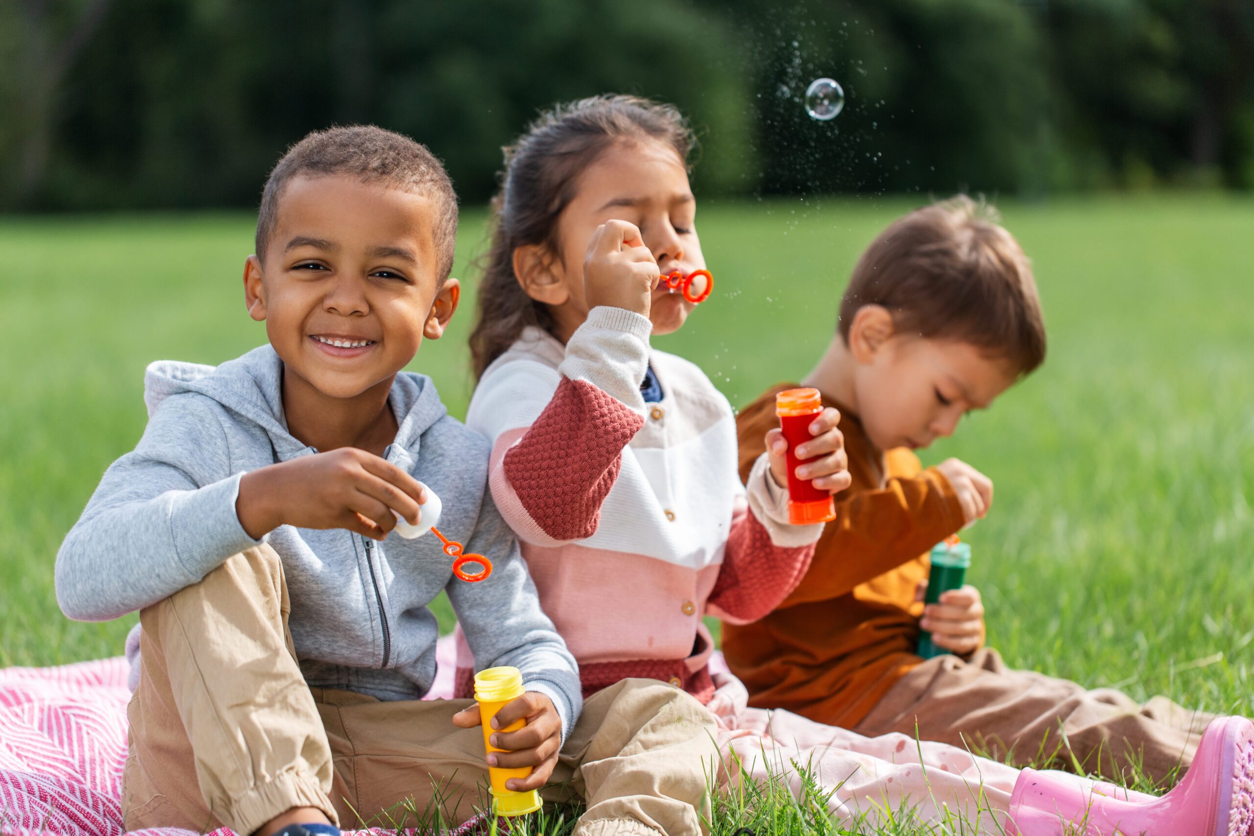 group of children, sitting on a blanket, blowing soap bubbles at park