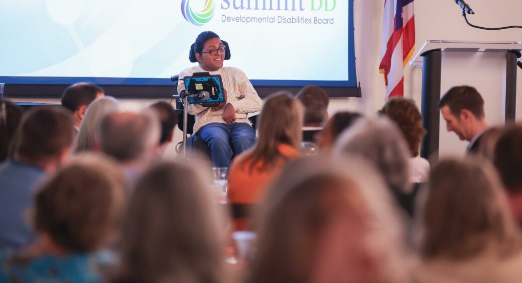 Young man in a wheelchair doing a keynote presentation on stage to a large, seated audience