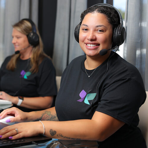 Woman sitting at desk smiling. Wearing a headset while using a laptop for remote monitoring services.