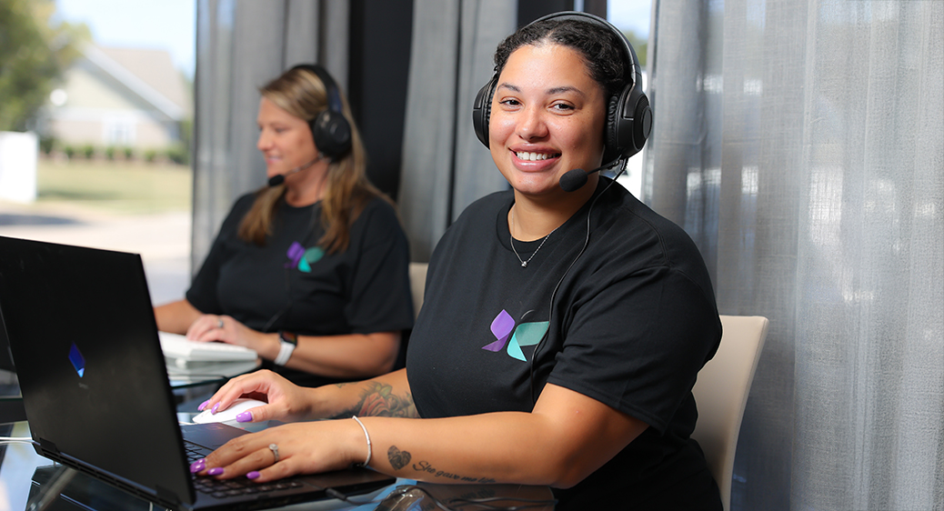 Woman sitting at desk smiling. Wearing a headset while using a laptop for remote monitoring services.