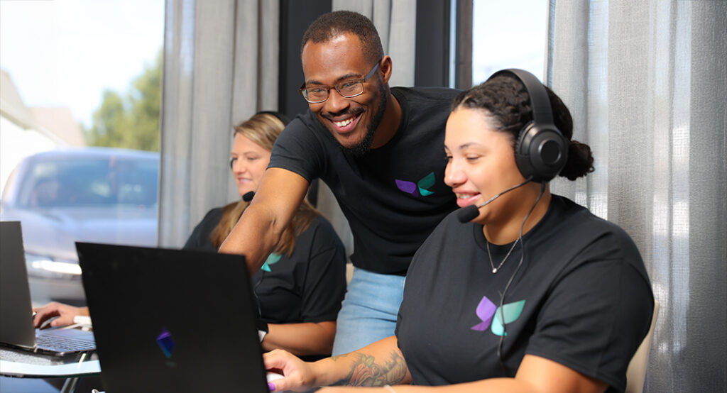 Woman and man sitting at desk smiling. Wearing a headset while using a laptop for remote monitoring services.