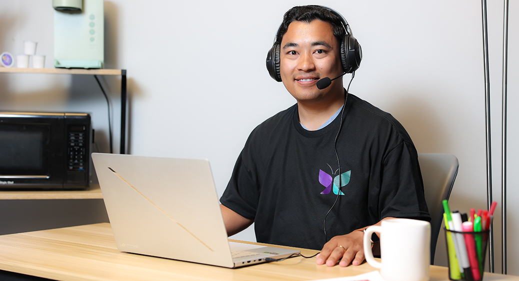 Man sitting at desk smiling. Wearing a headset while using a laptop for remote monitoring services.