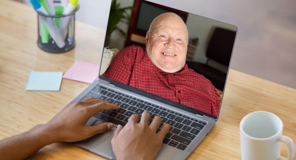 Older man on a laptop screen smiling in his living room while using an iPad for remote tech monitoring services.