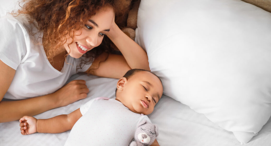 Young mom smiling next to her sleeping baby on a bed