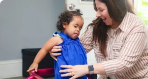 Smiling female developmental specialist helping young girl in a blue dress to stand.