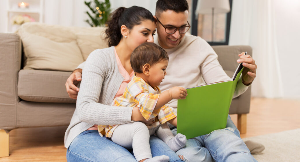 Mom, dad and baby sitting on the floor near couch reading a book to their toddler daughter