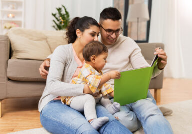 Mom, dad and baby sitting on the floor near couch reading a book to their toddler daughter