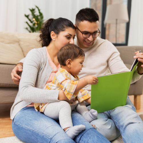 Mom, dad and baby sitting on the floor near couch reading a book to their toddler daughter