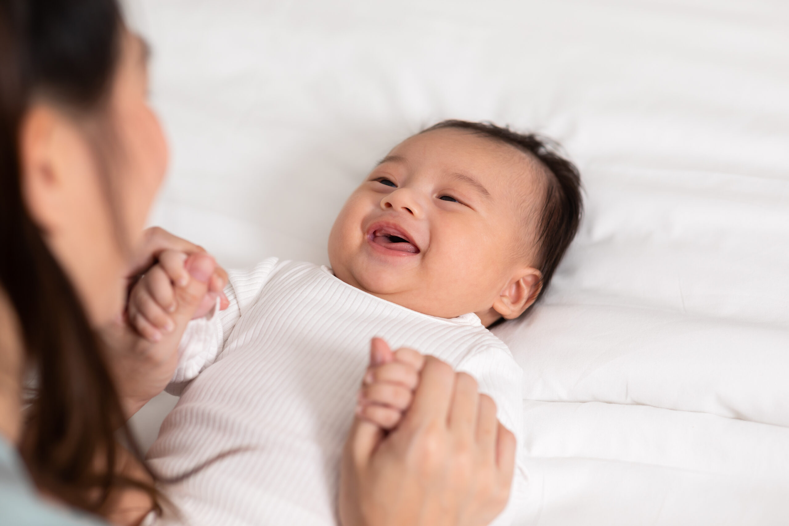 New asian mom playing to adorable newborn baby on bed smiling and happiness at home.Mom talking with infant baby and holding her hands laughing together.Baby and Mother day concept