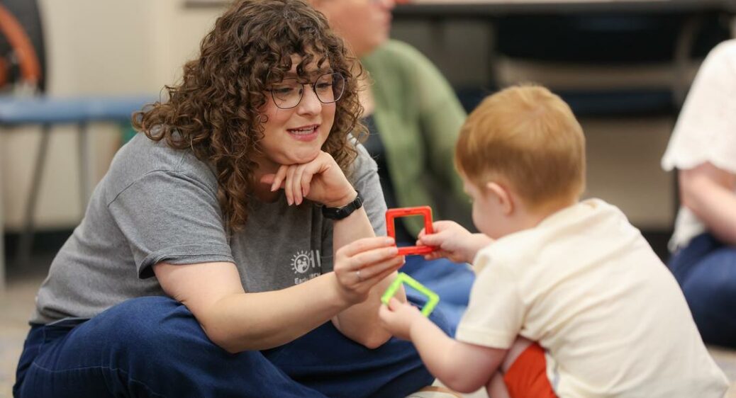 Young Speech Language Pathologist sitting on the floor hands a baby a red. plastic ring toy to hold
