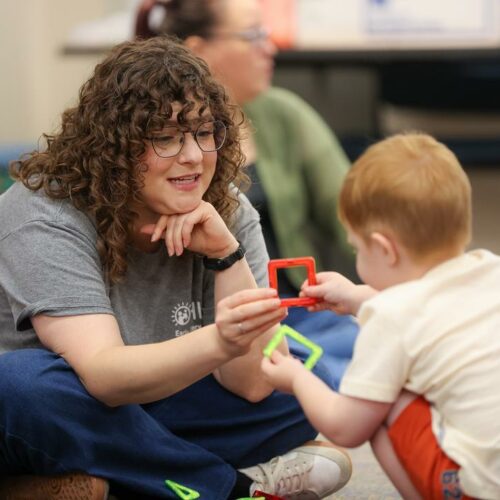 Young Speech Language Pathologist sitting on the floor hands a baby a red. plastic ring toy to hold