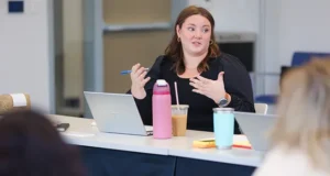 Image of Caucasian woman sitting at a table with her laptop in a discussion with another person beyond the camera angle