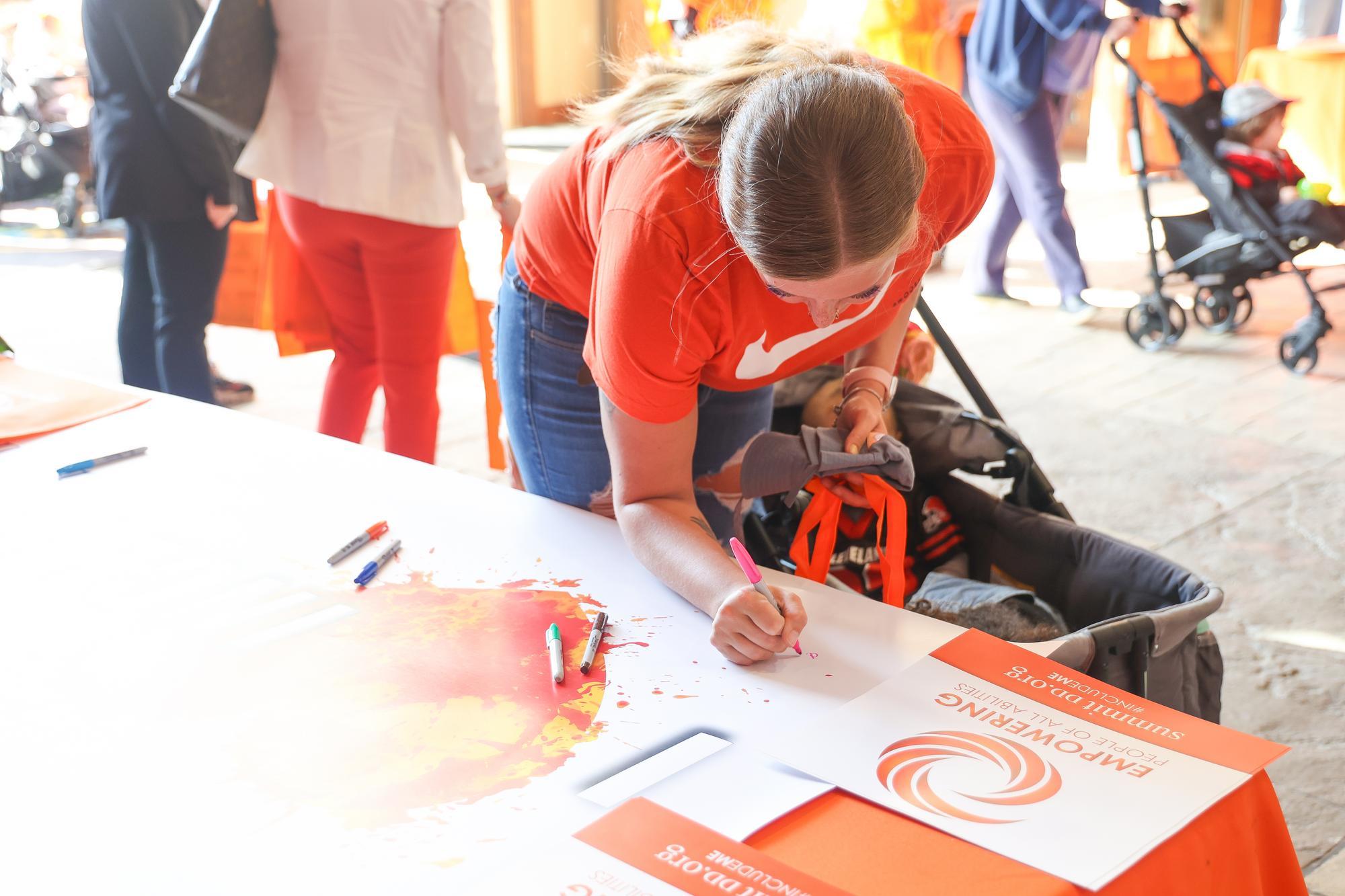 Woman in an orange shirt leaned over signing an "I heart Inclusion" banner