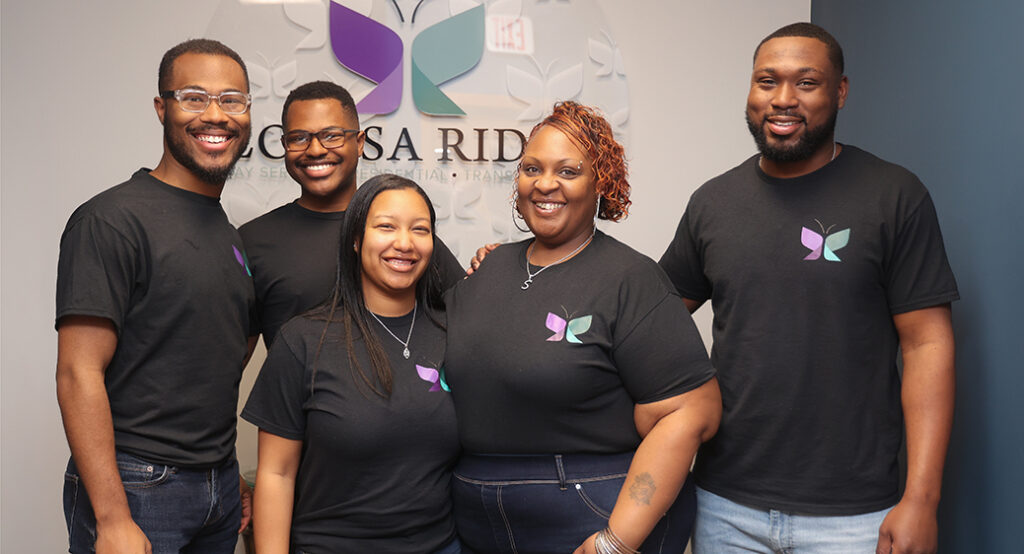 Smiling group photo of Louisa Ridge direct support professional team members. Standing in their lobby in front of a large Louisa Ridge butterfly logo.